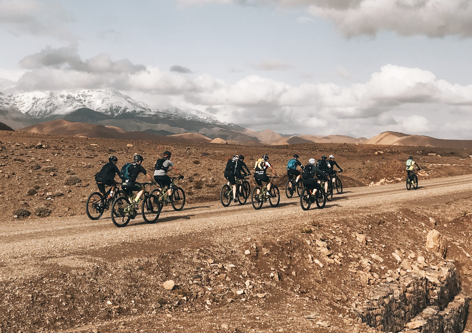 A group of riders on MTBs and E-Bikes on a wide track, facing the snowy peaks of the Atlas, Zagoride trip in Morocco with Exoride